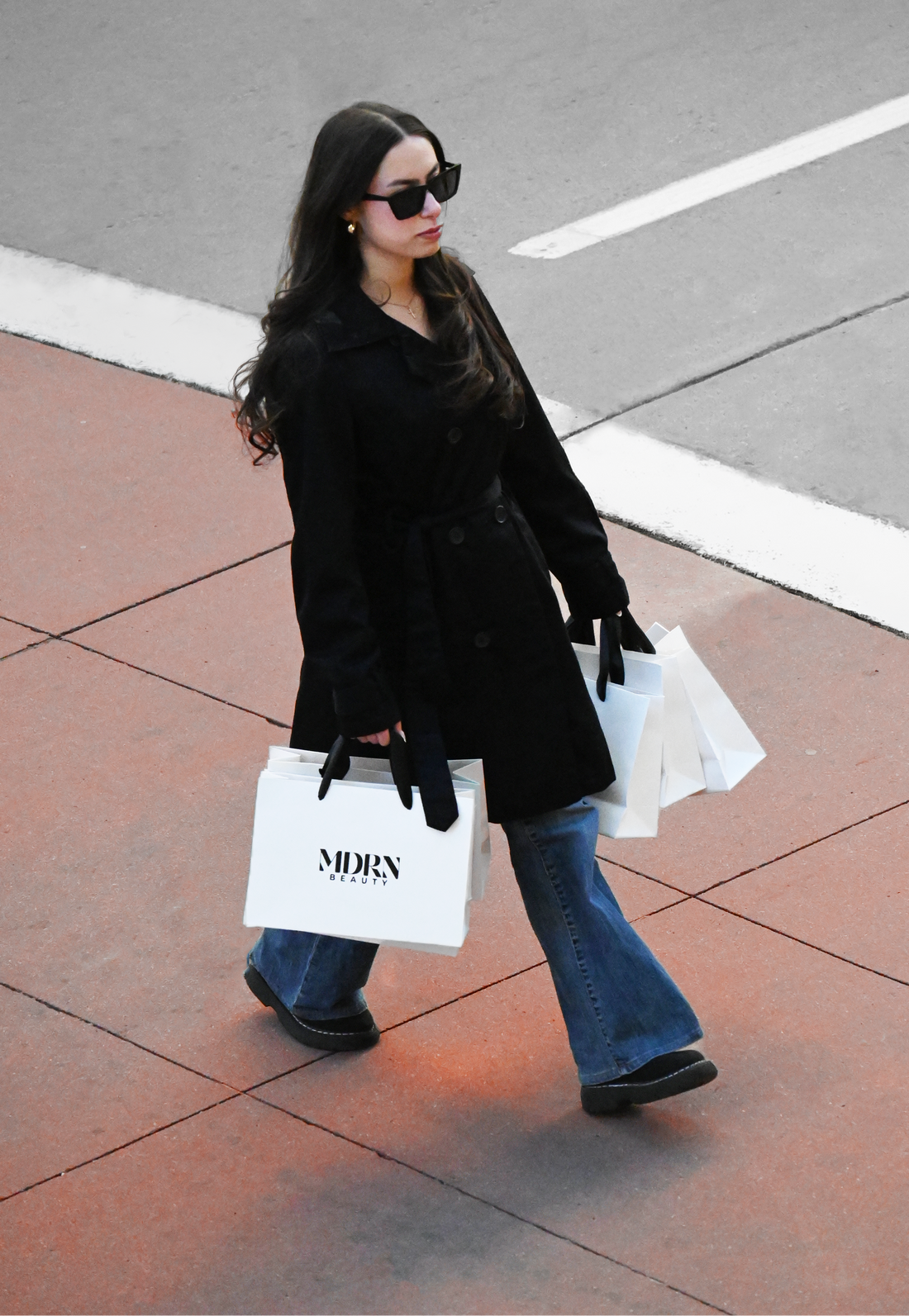 Woman in black coat and jeans holding shopping bags on a sidewalk.